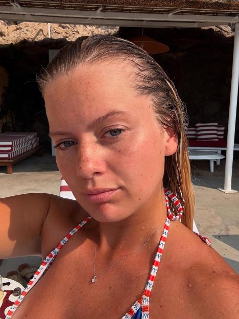 Close-up selfie of a woman with wet hair and freckles, wearing a red, white, and blue patterned bikini top.