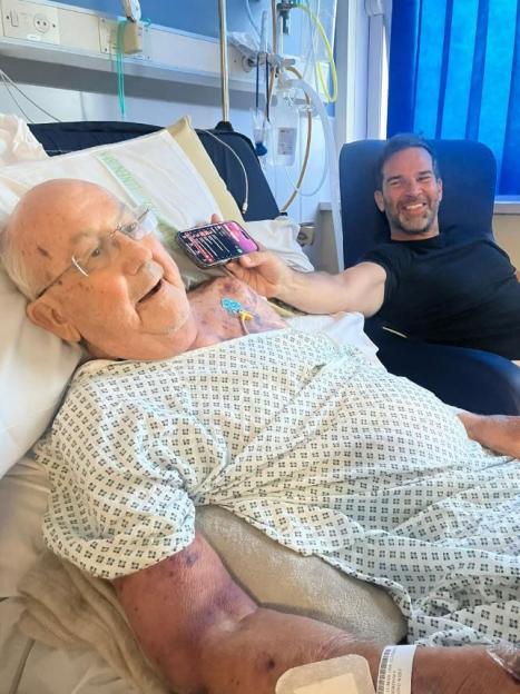 Elderly man in a hospital bed with a younger man sitting beside him.