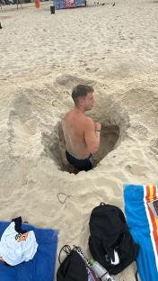 Man sitting in a hole in the sand on a beach.