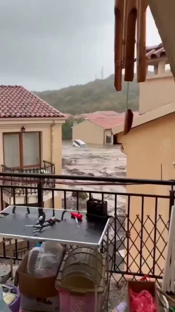 A flooded neighborhood with houses submerged in rushing water, viewed from a balcony.