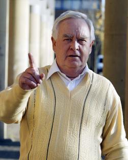 Older man in a tan sweater gesturing with his index finger.