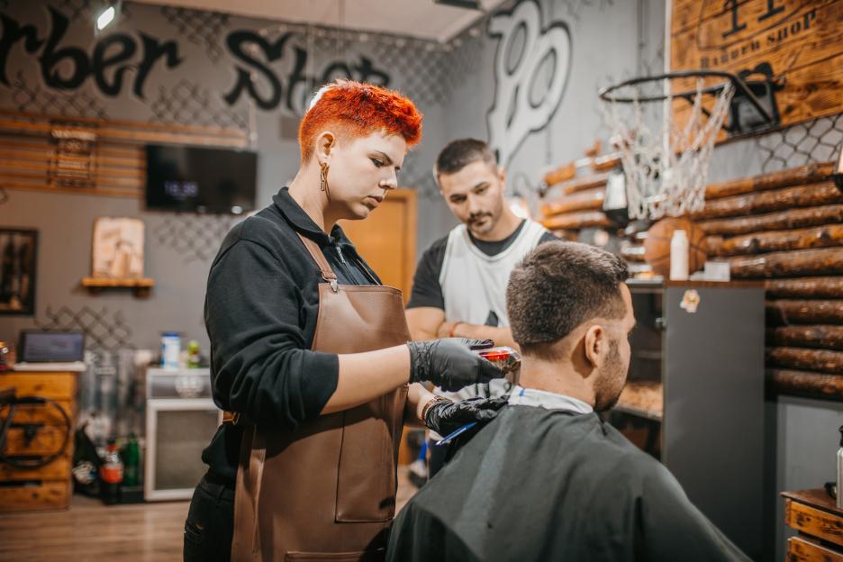 A young woman with short red hair cuts a man's hair in a barbershop, while another man observes.