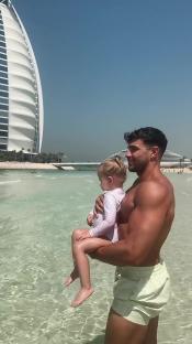 Man holding a toddler in shallow water with a tall building in the background.