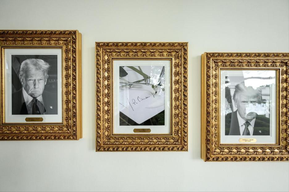 Three gold-framed pictures hanging on a white wall, including two black-and-white portraits of Donald Trump and a photo of an autopen signing "J.R. Biden."