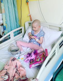 A young girl with no hair sits in a hospital bed, playing with toys.