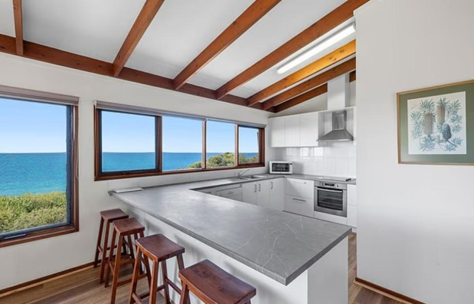 A kitchen with a long grey counter with four bar stools in the foreground, with windows overlooking the ocean in the background.