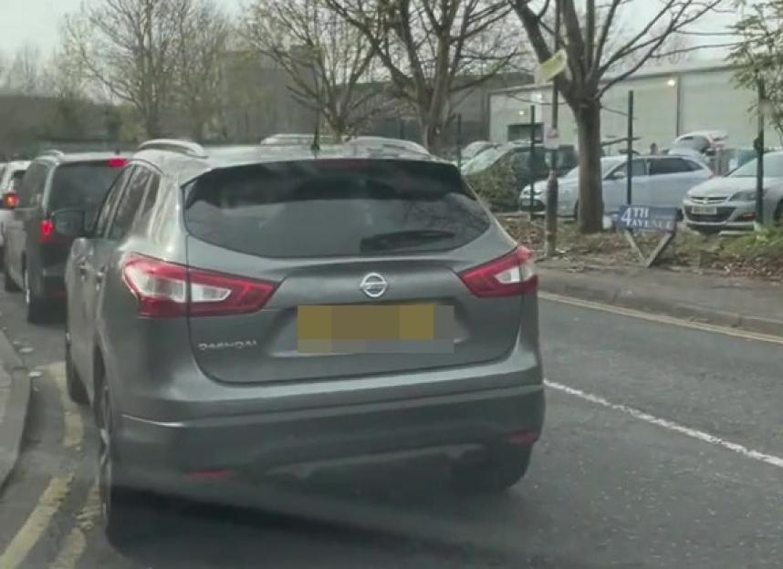 Cars lined up on a street with bare trees overhead, indicating a traffic queue.
