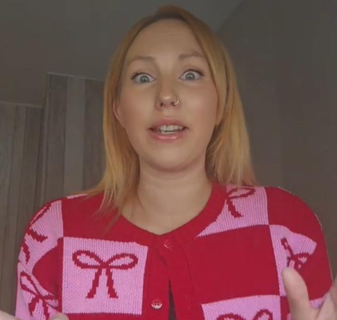 A woman with blonde hair and a nose piercing wearing a red and pink bow-patterned cardigan, looking forward with wide eyes.