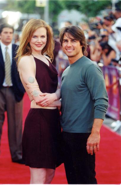 Nicole Kidman in a red mini-dress with a gold arm cuff and Tom Cruise in a gray long-sleeved shirt, both smiling on a red carpet.