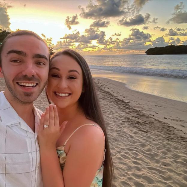 Nathan Sykes and Charlotte Burke smiling with an engagement ring visible on a beach at sunset.