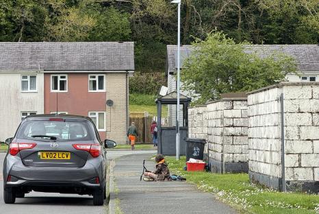 A child sits on a sidewalk with a scooter, near houses and a parked car.