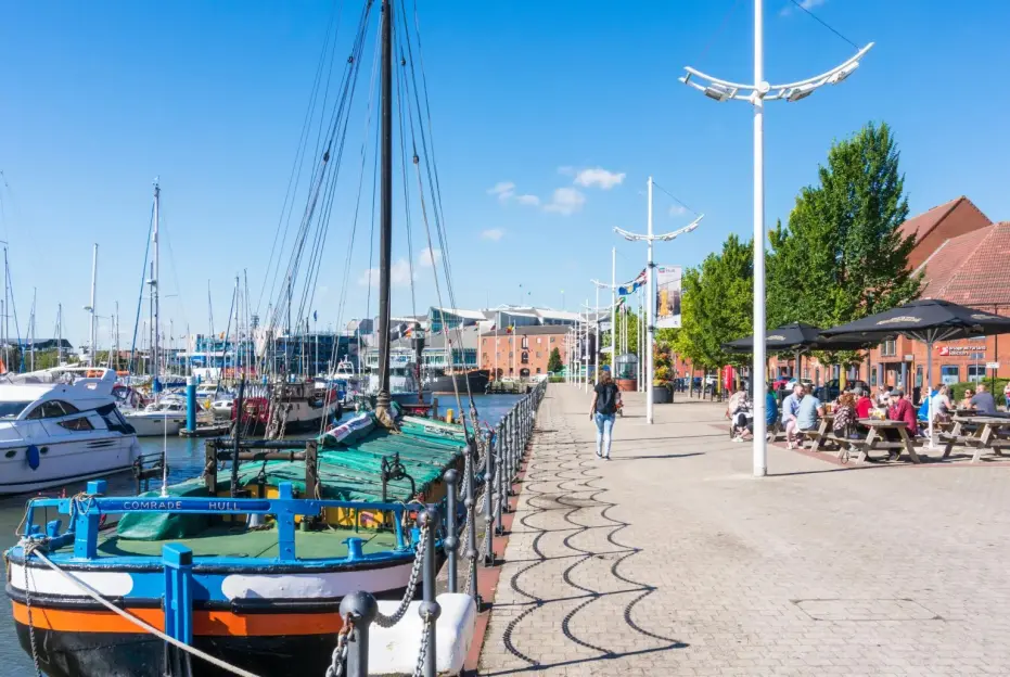 People eating and drinking at outdoor tables on a sunny day at a marina with boats and buildings.