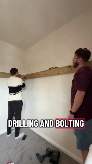 Two men drilling and bolting wooden planks to a white wall in a room with gray carpet.