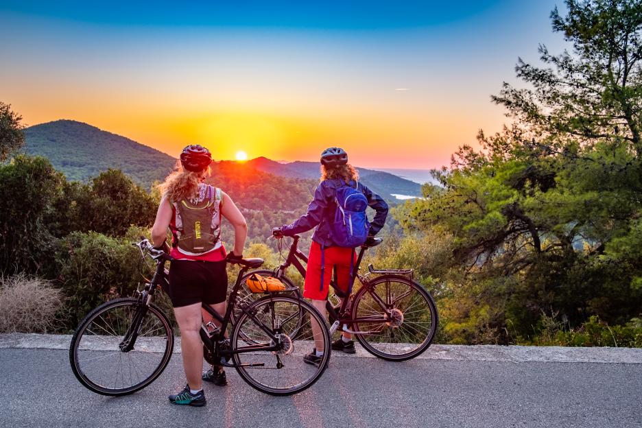 Two people cycling while watching a sunset over mountains and the sea.