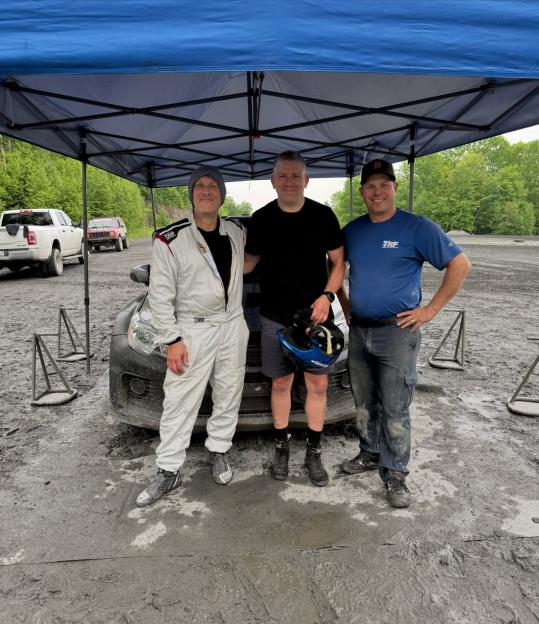 Three men stand under a blue tent, one in a white racing suit, next to a black car on a muddy track.