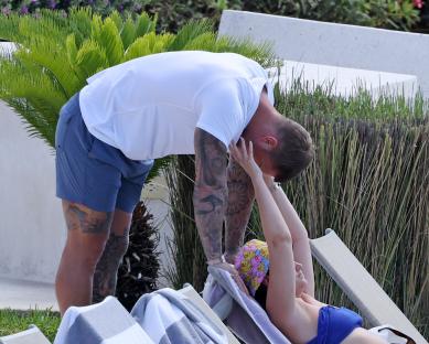 A man leans down to kiss a woman relaxing on a lounge chair by a pool.
