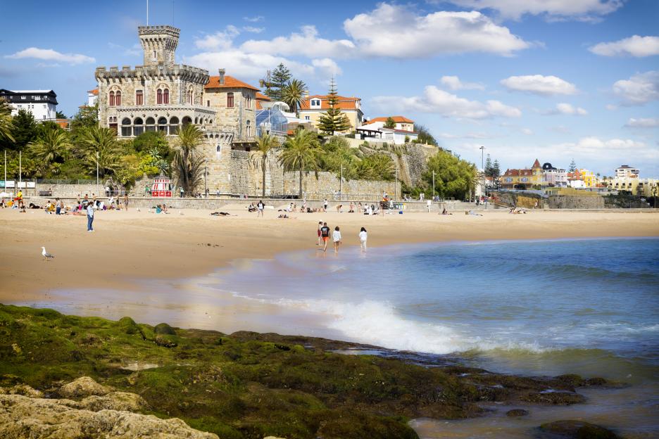 Tamariz beach in Estoril, Portugal, with a historic stone building and palm trees overlooking the sandy shore where people are relaxing and walking by the Atlantic Ocean.