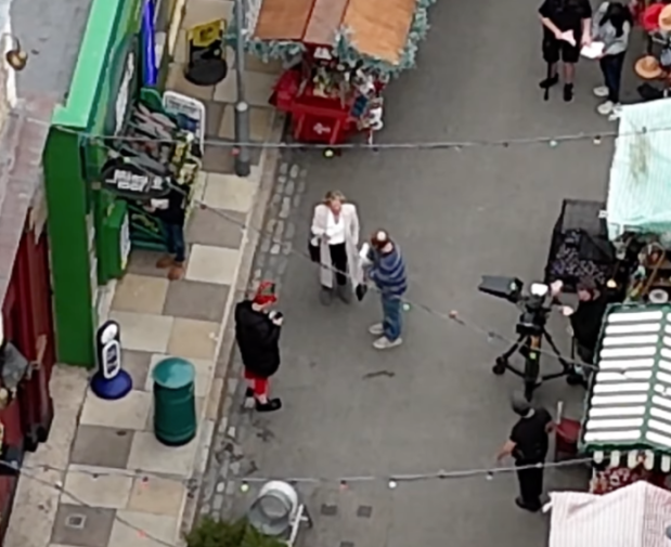An overhead view of a film crew interviewing people on a street lined with shops and market stalls.