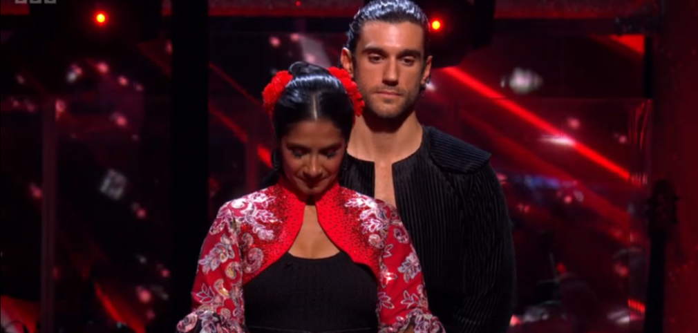 A woman in a red and white floral bolero and a man in a black shirt stand on a stage with red lighting.
