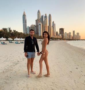Couple holding hands on a beach with a city skyline in the background.