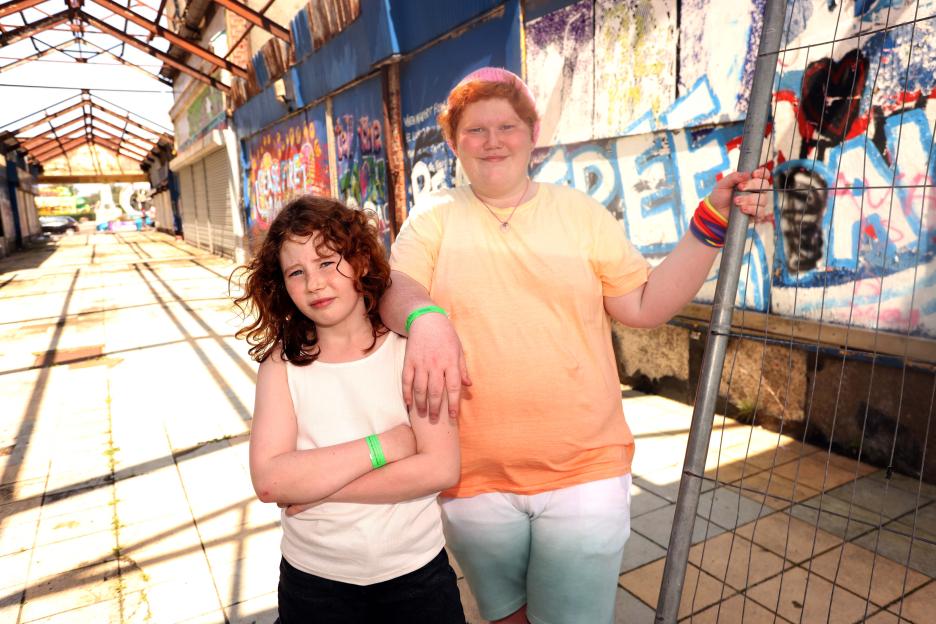 Two children stand in front of a graffiti-covered wall in a dilapidated arcade.