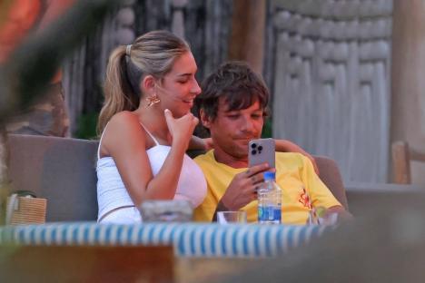 Louis Tomlinson and Zara McDermott looking at a phone together at a beachside restaurant.
