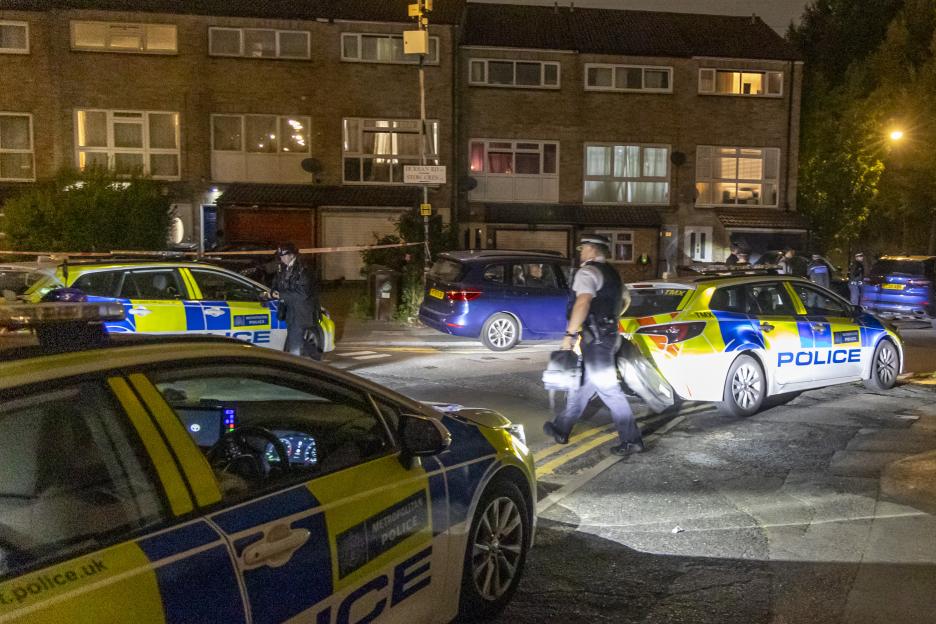 Police officers and marked police cars at the scene of a shooting in Walthamstow, London, at night.