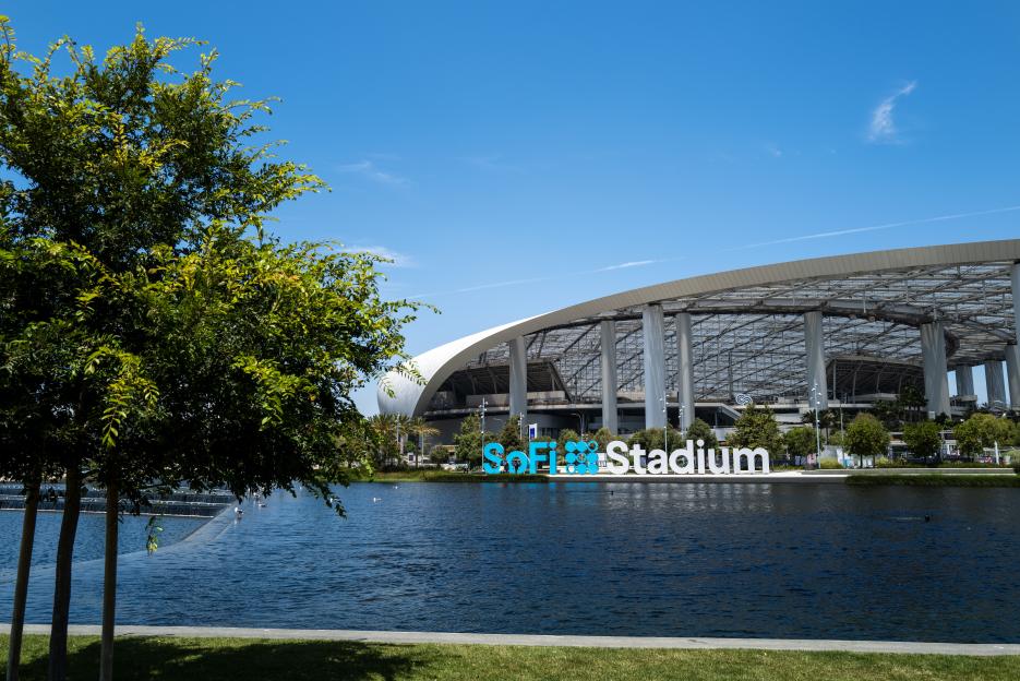 View of SoFi Stadium and the SoFi Stadium sign across a body of water, framed by green trees and grass.