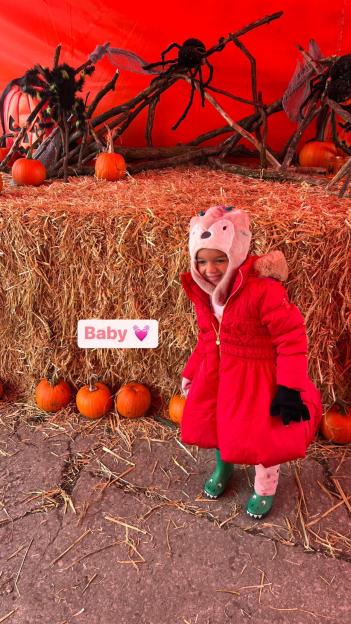 A young child in a red coat and animal-eared hat stands in front of hay bales and pumpkins, with spider and pumpkin decorations in the background.