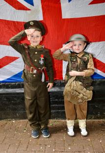 Two young boys in World War II costumes salute in front of a Union Jack flag.
