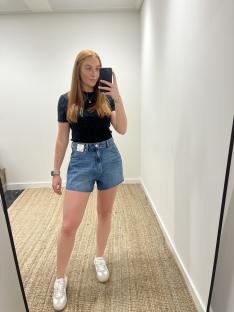 Woman in denim shorts and a black shirt taking a selfie in a mirror.
