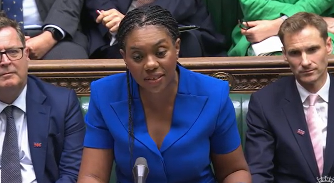 Woman in blue speaking at a podium flanked by two men in suits.