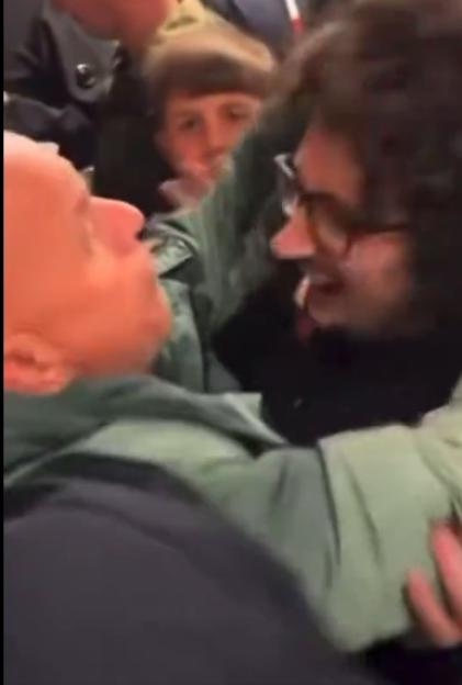 A man with short hair and a man with curly hair and glasses hug.
