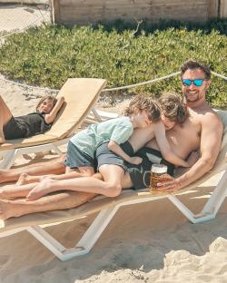 Man relaxing on a beach lounger with his two young sons asleep on him, holding a beer.