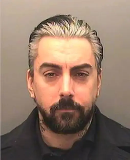 Mugshot of a man with graying black hair, a beard, and neck tattoos.