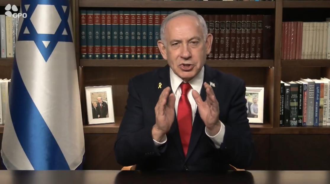 Benjamin Netanyahu addressing the camera, with an Israeli flag and bookshelves in the background.