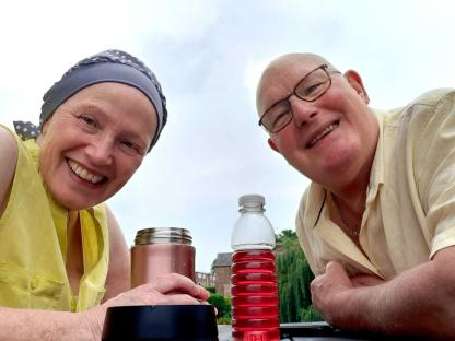 A bald couple smiles for a selfie outdoors with drinks.