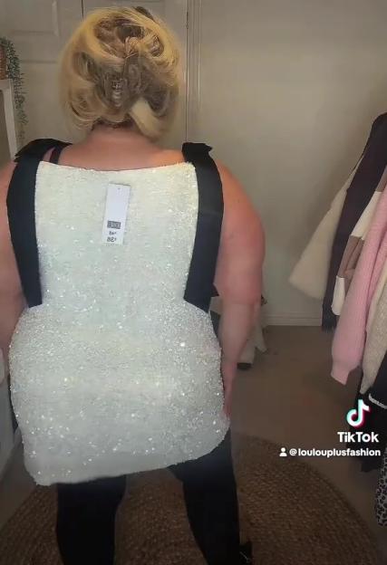 Rear view of a plus-size woman trying on a white sequined mini dress with black bow shoulder straps.