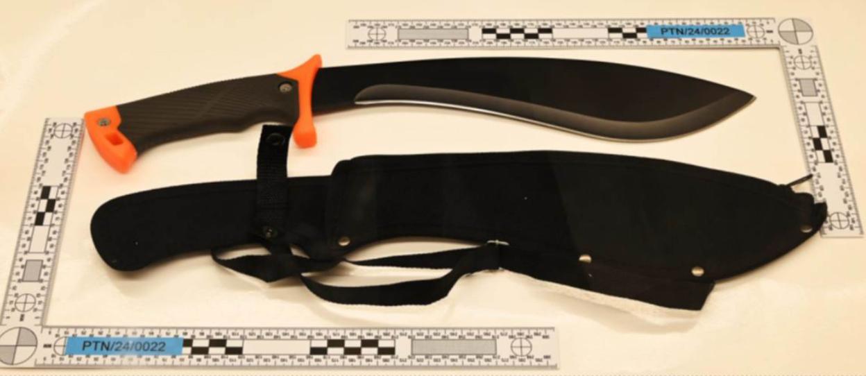 A large knife with an orange and black handle, a black blade, and a black sheath lying on a white surface with crime scene measuring rulers.