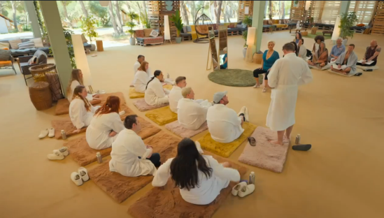 Group of people in white robes sitting on mats in a large room.