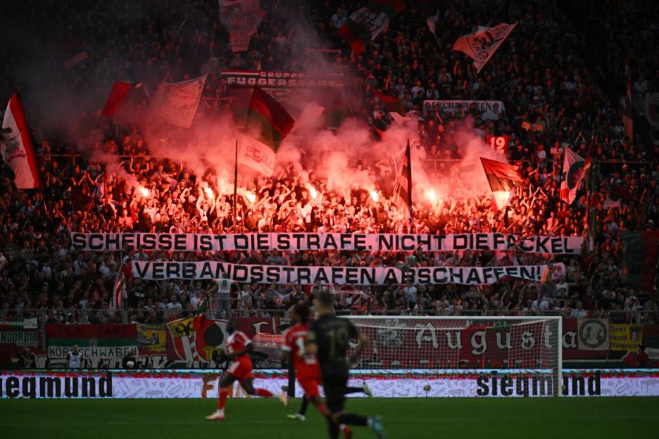 Soccer fans in a stadium holding flares and a banner that reads "Scheisse ist die Strafe, nicht die Fackel Verbandsstrafen abschaffen!"