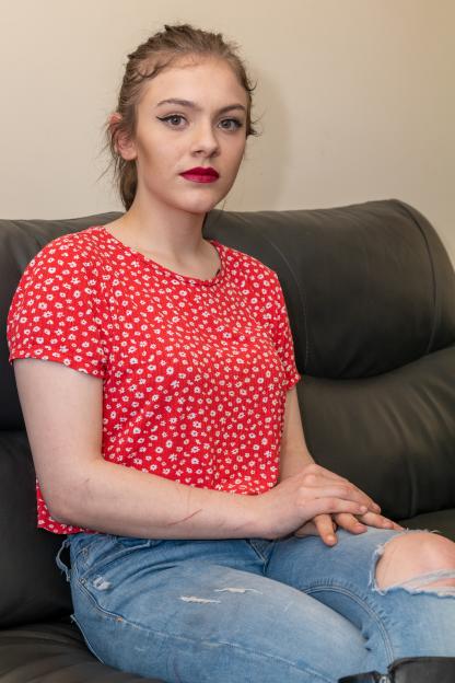 Rebekah Elizabeth Simpson, wearing a red floral top and ripped blue jeans, sits on a black couch.