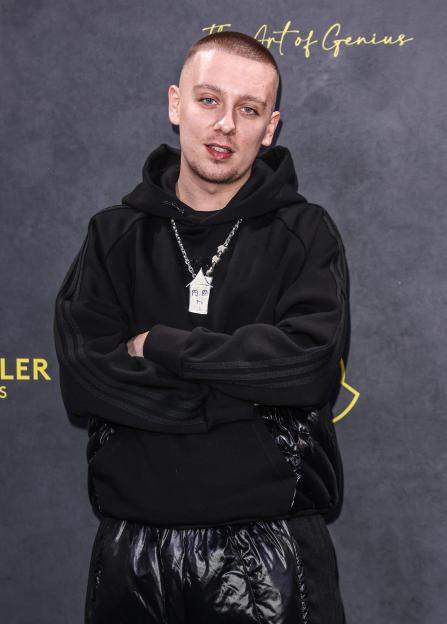 Aitch at the Moncler Genius show wearing a black hoodie and shiny black pants, accessorized with a house-shaped pendant.