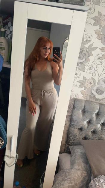 A woman with long red hair and a beige two-piece outfit takes a mirror selfie while holding a phone.
