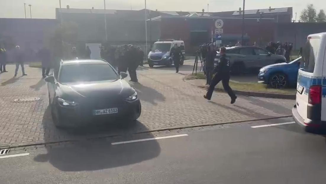 A dark car with German license plates HH-WZ 8103 driving on a paved road, with multiple people, including police officers, and other vehicles in the background.