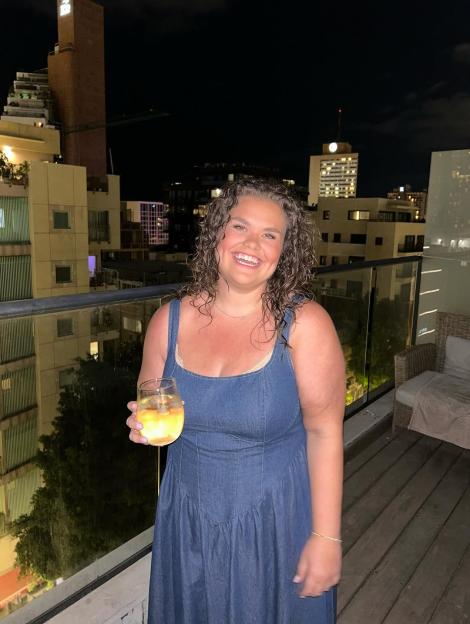 Amy Tapper smiling in a denim dress while holding a drink on a balcony at night.