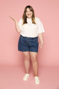 Woman in white shirt and denim shorts gesturing against pink background.