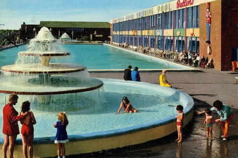 A large outdoor swimming pool at a Butlin's resort, with a fountain in the foreground and people relaxing by the pool.
