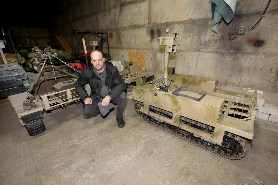 A man in a black coat kneels between two Unmanned Ground Vehicles (UGVs) in a workshop.