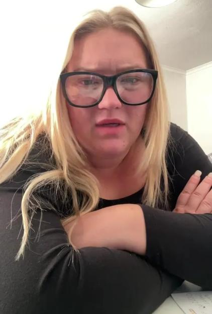 A blond woman wearing black glasses looks at the camera with a worried expression.